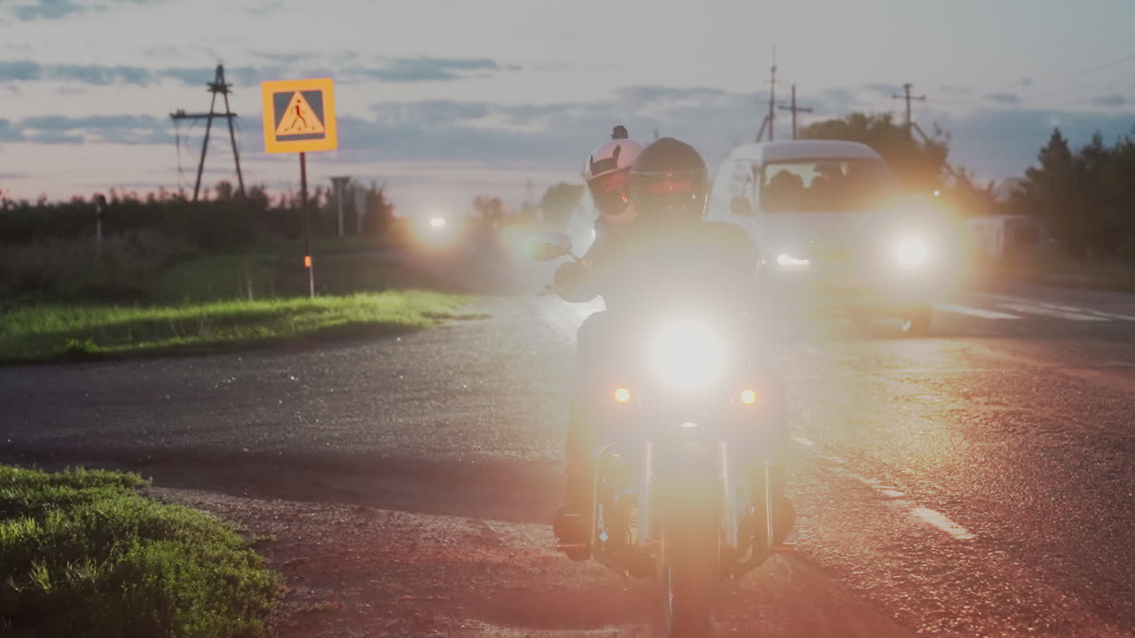 Biker wearing helmet pulls over along T junction under glowing headlight as cars drive past with headlights shining, pedestrian road sign visible by roadside and evening sky filled with clouds
