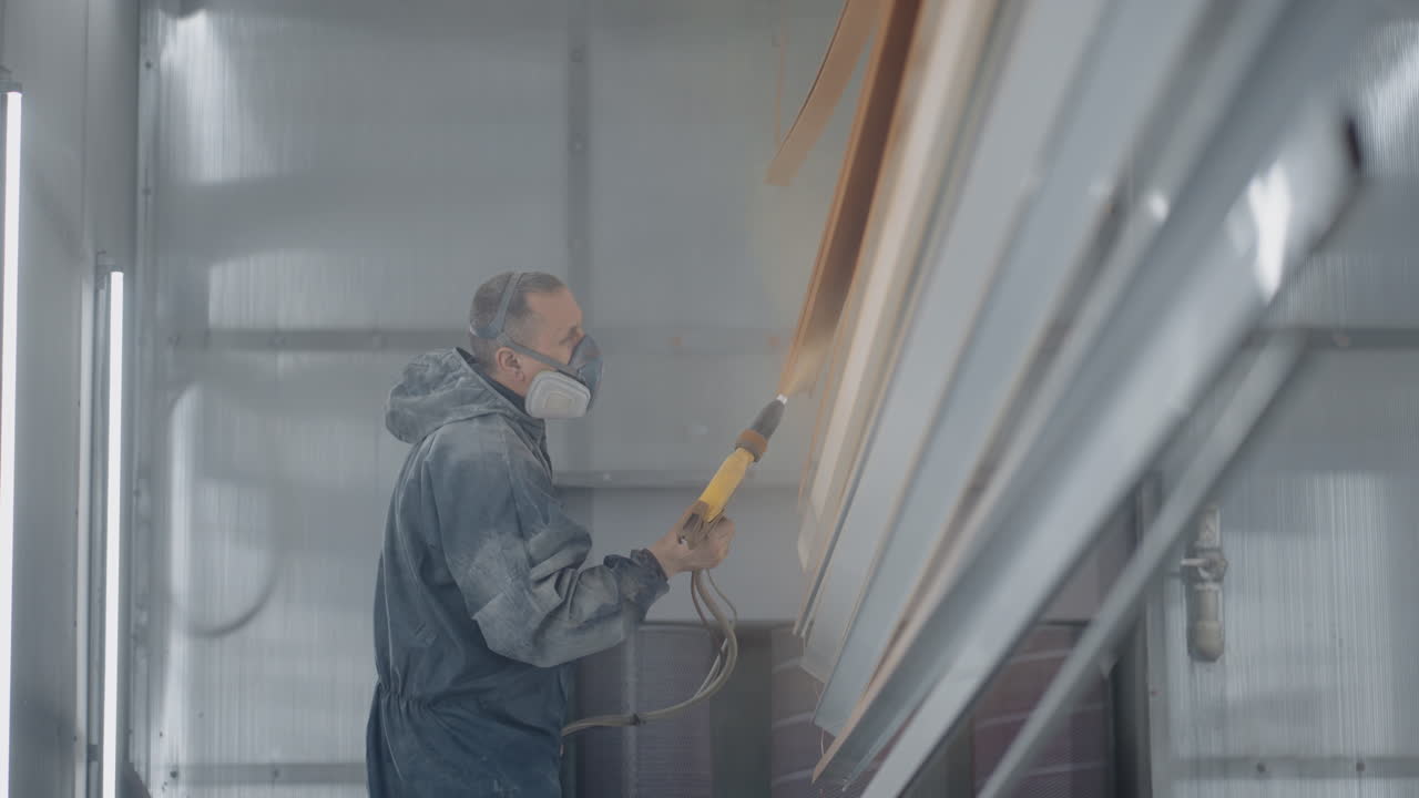 A man in a protective suit and mask sprays paint through a spray gun in slow motion on steel parts.
