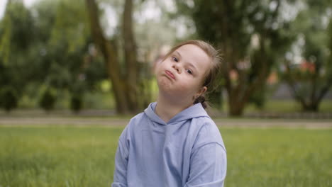 Kleines-Mädchen-Mit-Down-Syndrom,-Das-In-Die-Kamera-Lächelt-Und-Auf-Dem-Gras-Im-Park-Sitzt