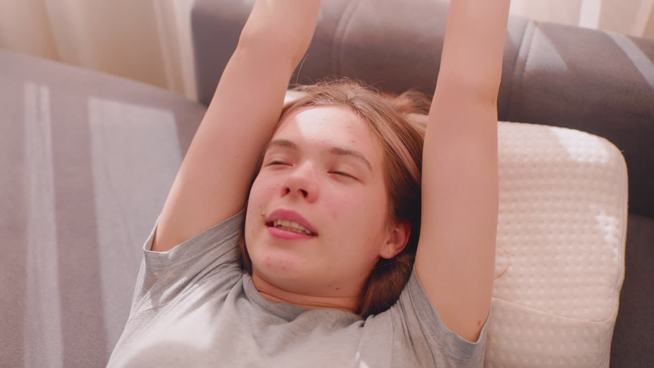 Close up of young woman lying on couch pillow slowly waking up after long sleep, stretching arm in soft daylight, relaxed expression reflecting peaceful rest and refreshed morning mood
