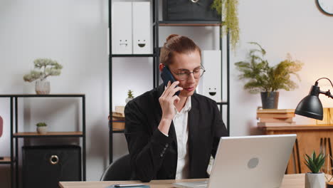 hombre de negocios caucásico elegante comunicándose con el cliente por teléfono inteligente y usando una computadora portátil en el escritorio