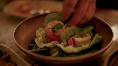 a hand arranges shrimp and greens on a plate under soft, warm lighting, creating an inviting culinary scene