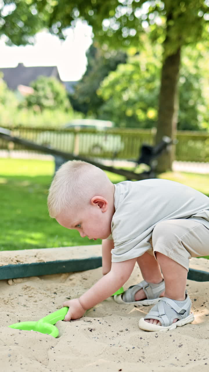 Un bambino piccolo in maglietta e pantaloncini durante una passeggiata al parco giochi. Il bambino gioca nella sabbiera, toccando la sabbia con le manine. Il bimbo scava la sabbia con una paletta.