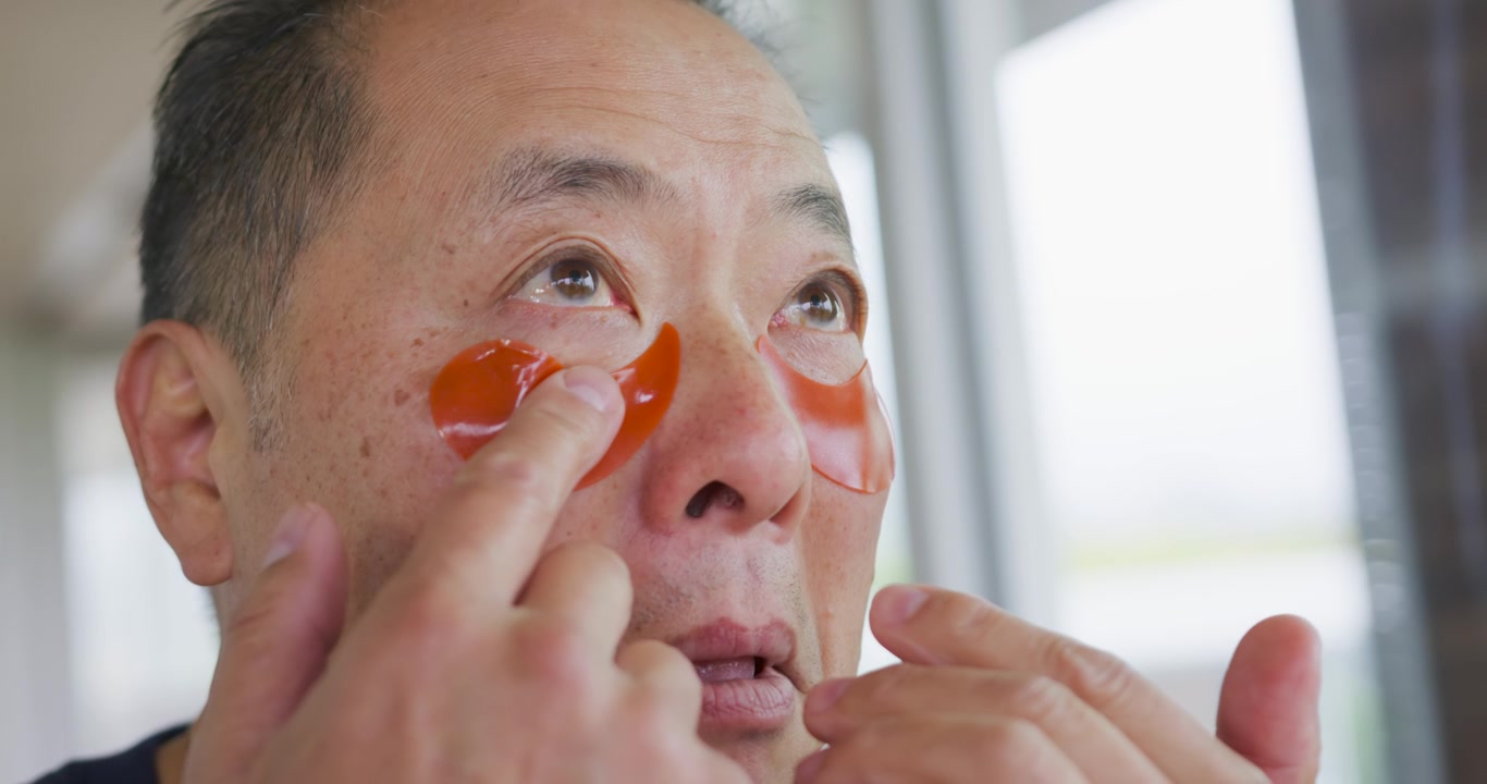 Senior Chinese man bringing orange eye patches applying pressing for care at home by sunny window