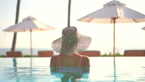 a woman wearing a straw sun hat, while relaxing in a resort, swimming pool looks out the ocean beyond