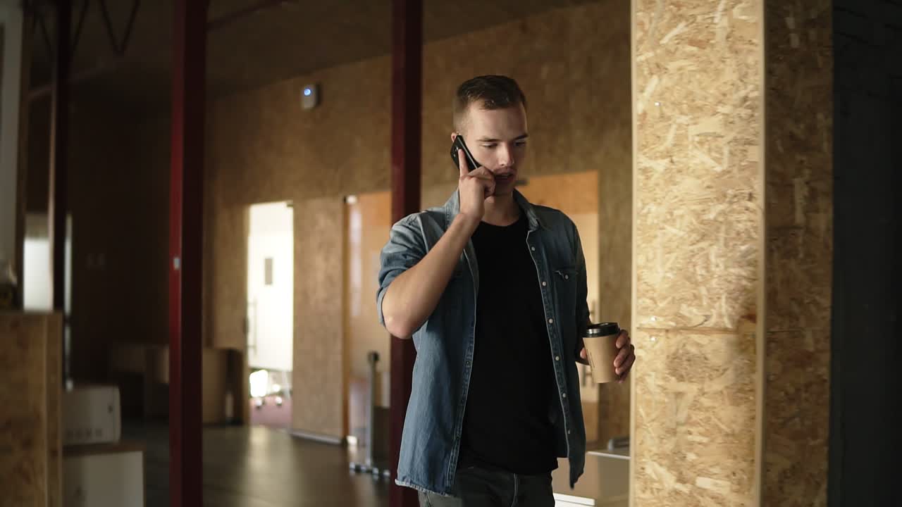 Young Man In Casual Walks At A Modern Office Or Workplace With Brick Style Interior While Talking By His Mobile Phone