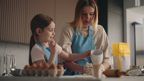 cute little daughter is helping to her mother in kitchen pouring water on dough woman is kneading by hands