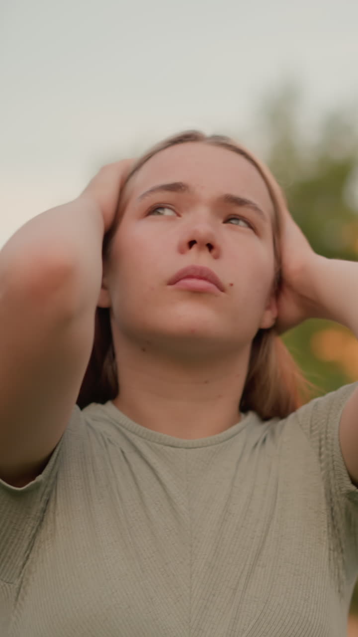 joven al aire libre con las manos presionadas contra su cabeza, los ojos cerrados, emanando una expresión contemplativa y de mal humor, rodeada de elementos urbanos y verdes borrosos