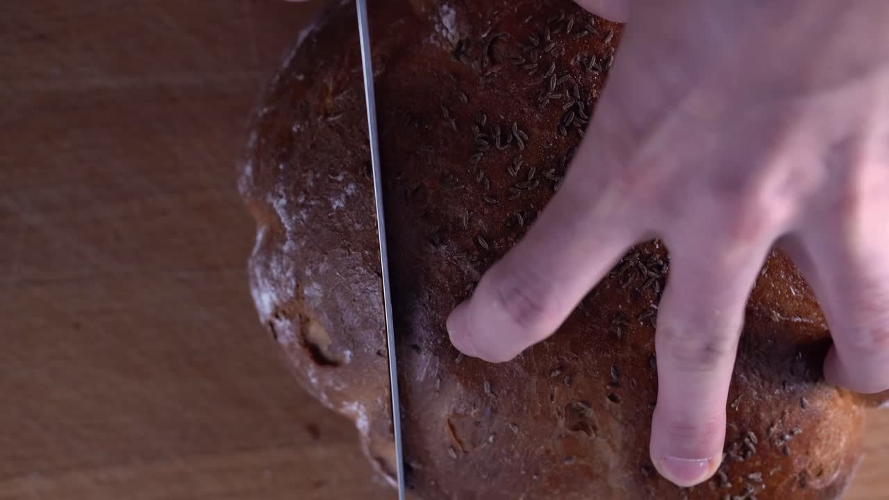 Long serrated blade of knife slicing through the crust of fennel seed bread on wooden chopping board with black background, SLOW MOTION, OVER THE TOP VIEW