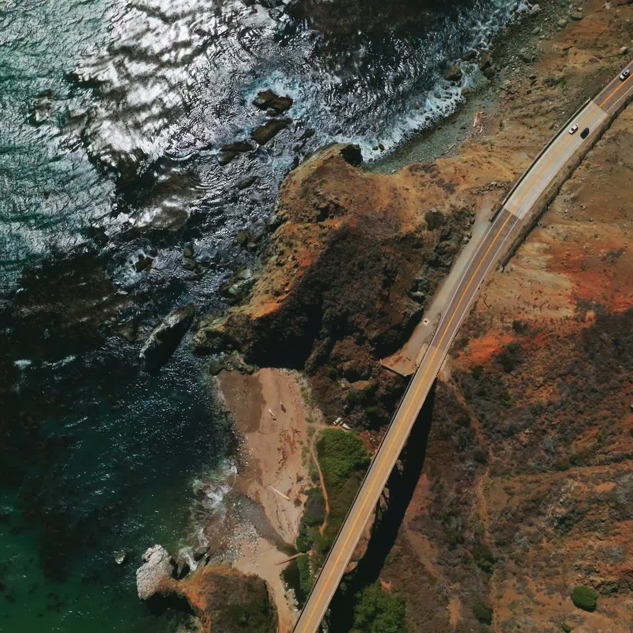View of the roads in the mountains connected by the bridge. Rocky coastline of California with beautiful blue ocean from aerial perspective