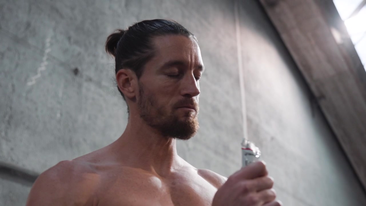 Un'inquadratura dal basso di un giovane muscoloso a torso nudo con i capelli legati all'indietro e la barba, che indossa un orologio al polso destro e mangia un gel energetico sportivo