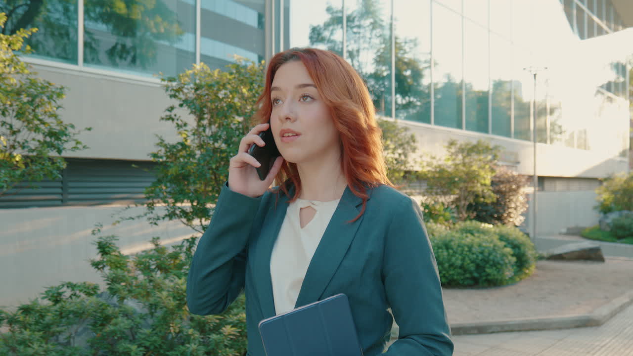 Shot of caucasian businesswoman wearing formal suit walking though city sidewalk while talking phone conversation on the way to office