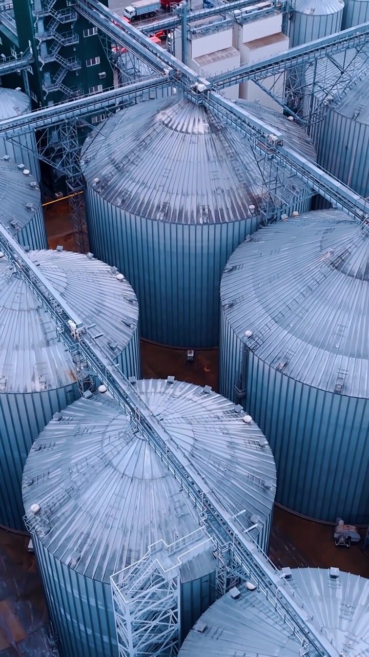 Ripresa dall'alto sopra le cime di enormi silos per cereali. Camion con prodotti agricoli arrivano nel territorio di uno stabilimento agricolo. Vista aerea. Video verticale