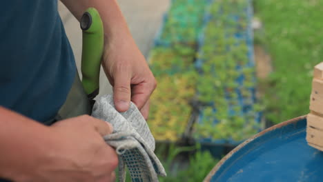 adult gardener in pocket apron picks up work gloves and tucks them into side pocket beside blue metal drum and wooden crate, close view of hands preparing for garden task