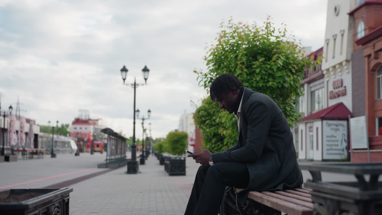 Una bonita calle de la ciudad rodeada de edificios modernos y árboles verdes, con un maletín en la mano y usando el teléfono, capturando un estilo de vida urbano tranquilo, un clima fresco y un entorno profesional al aire libre