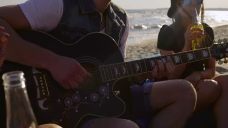 young happy friends drinking beer, sitting on easychairs on the beach and listening to a friend playing guitar on a summer evening. shot in 4k.