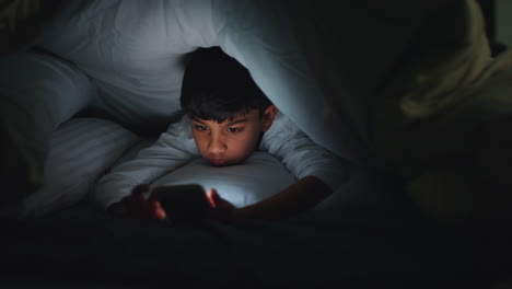 close up of young boy in bedroom at home using mobile phone to text message under covers or duvet at night 10