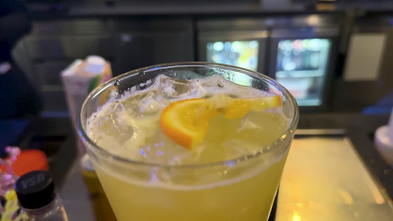 A bartender in formal attire prepares a citrus cocktail at a modern bar in Bangkok, under cool ambient lighting, with steady camera focus on the drink
