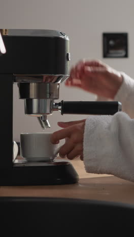 una mujer pone una taza en la bandeja de agua de la máquina de café en el primer plano de la cocina. una dama con ropa acogedora llena la taza con una bebida fuerte y caliente de la cafetera en casa. aparatos domésticos