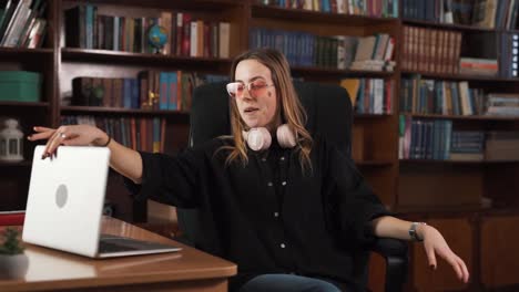 en el fondo, lleno de numerosos libros, una chica de contenido se sienta cómodamente en una silla, diligentemente ocupada con su computadora portátil