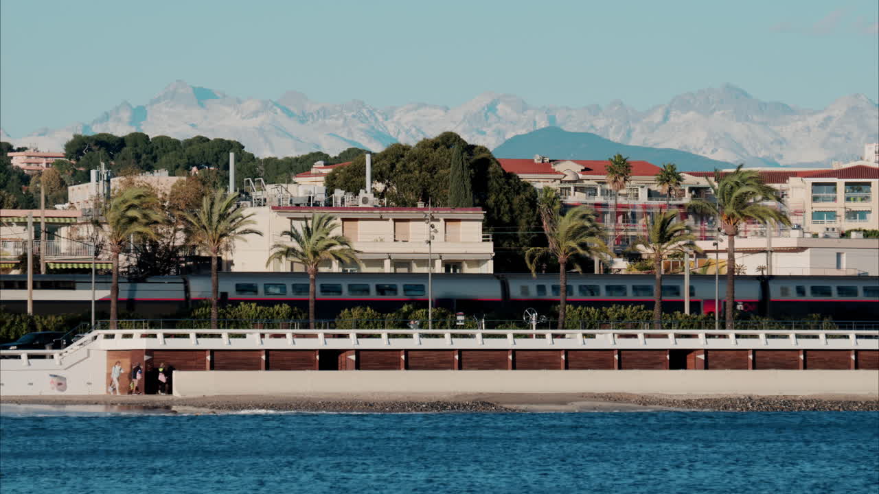 Train moving in front of buildings and palm trees on the shore with a view of the mountains in the background in Antibes, France