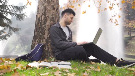 a young student sits leaning against a tree in the park and cheerfully works with a laptop.