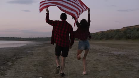 Young-woman-and-man-running-on-the-sand-with-raised-american-flag,-feeling-freedom.-Evening-dusk.-Tracking-footage