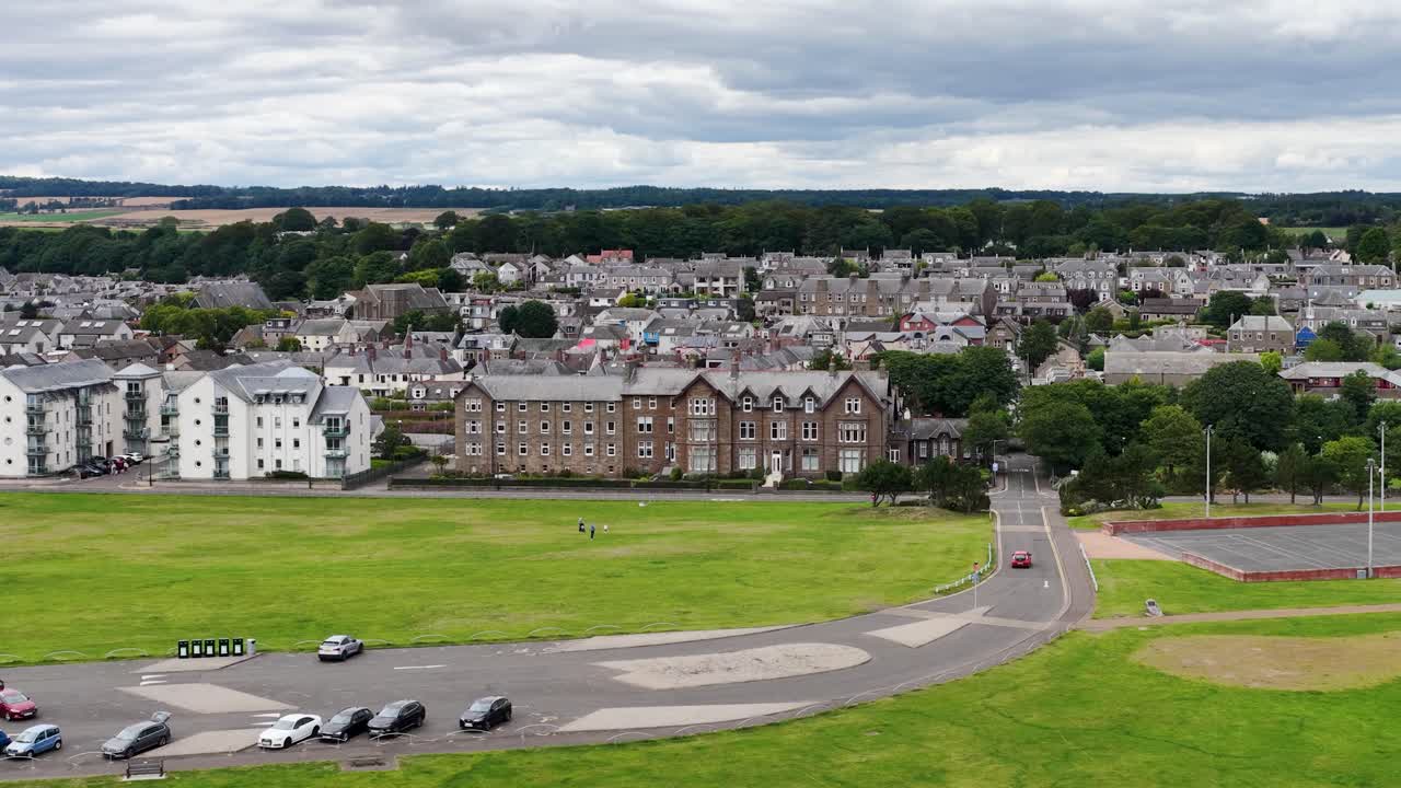 Drone ascends from beach over rocky seawall, revealing golf course, historic buildings, and townscape