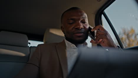 confident man businessman with black skin color talks on the phone and looks at his tablet to remember his plans while driving in a modern car in a non-urban area