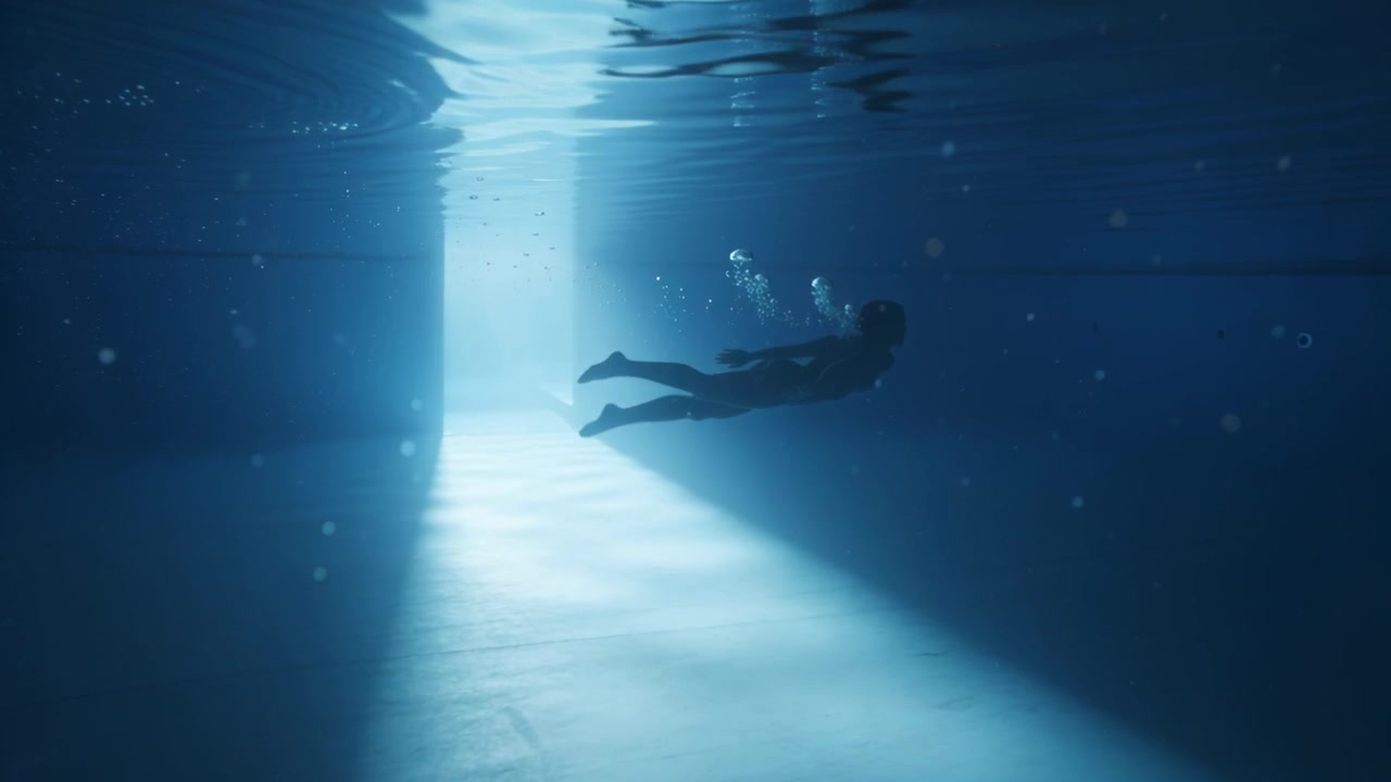 Nuotatrice che scivola in costume scuro in piscina, notando un fascio di luce, scalciando ed espirando bolle