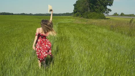 slow motion young girl runs across green field in red dress that flutters in the wind she takes off her hat