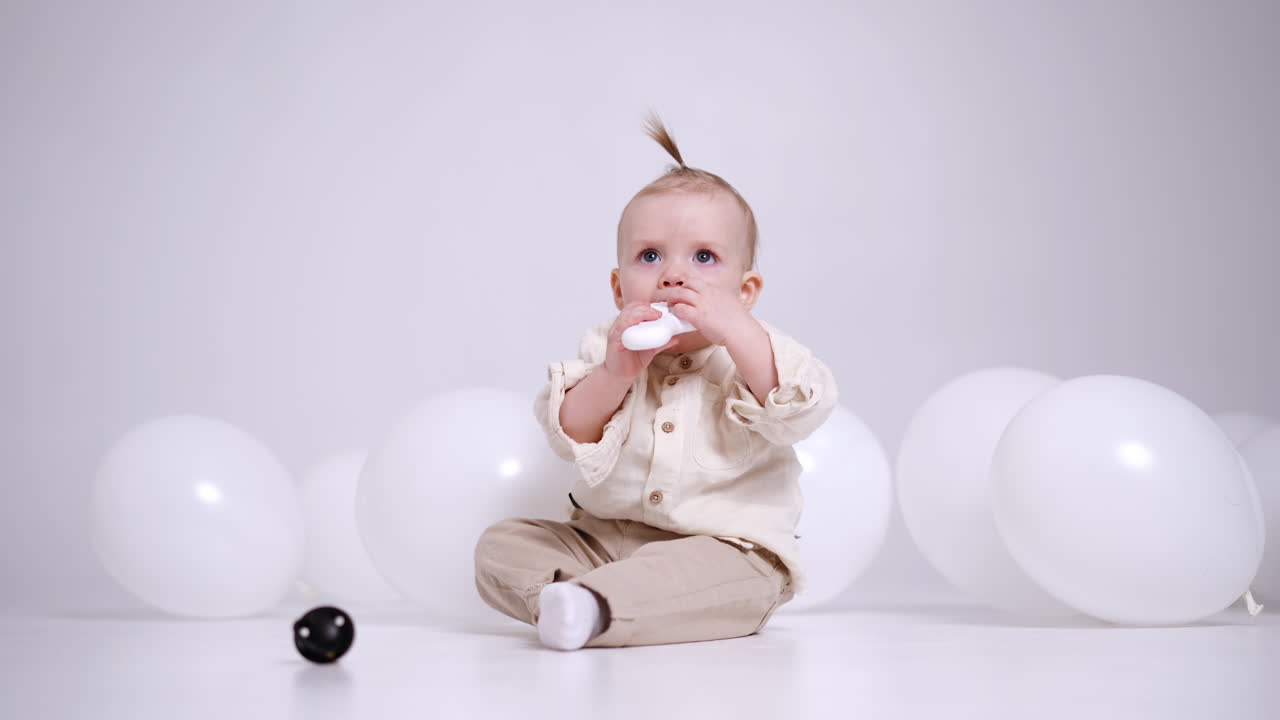 Little adorable Caucasian baby sits on the floor in studio among the white balloons. Cute child plays with toys and pulls it to mouth.