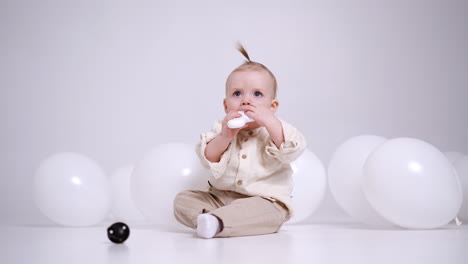 little adorable caucasian baby sits on the floor in studio among the white balloons. cute child plays with toys and pulls it to mouth.