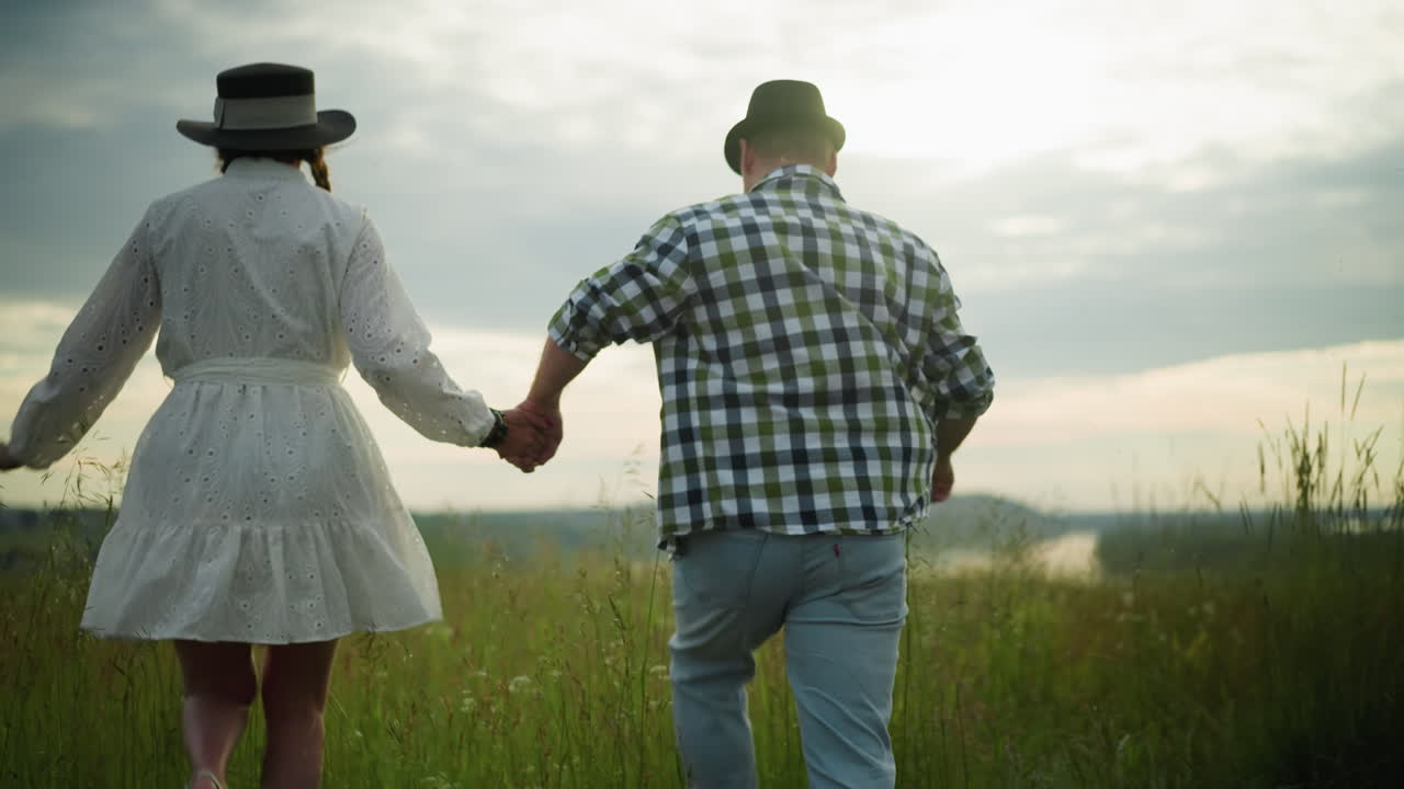 Back view of a couple holding hands and running through a grassy field at sunset. The man wears a black hat and checkered shirt, while the woman is dressed in a white dress and hat