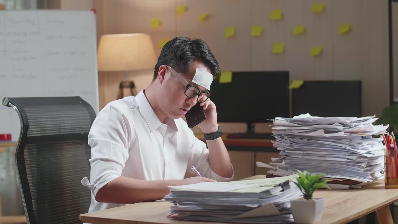 병든 아시아인 남자가 사무실에서 문서와 함께 열심히 일하면서 전화로 이야기하고 메모를 취하고 있습니다.