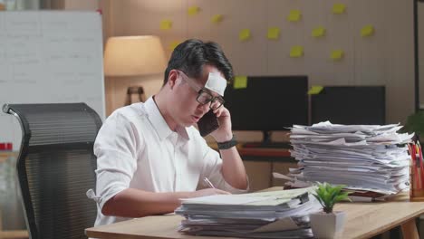 sick asian man talking on phone and taking note while working hard with documents at office