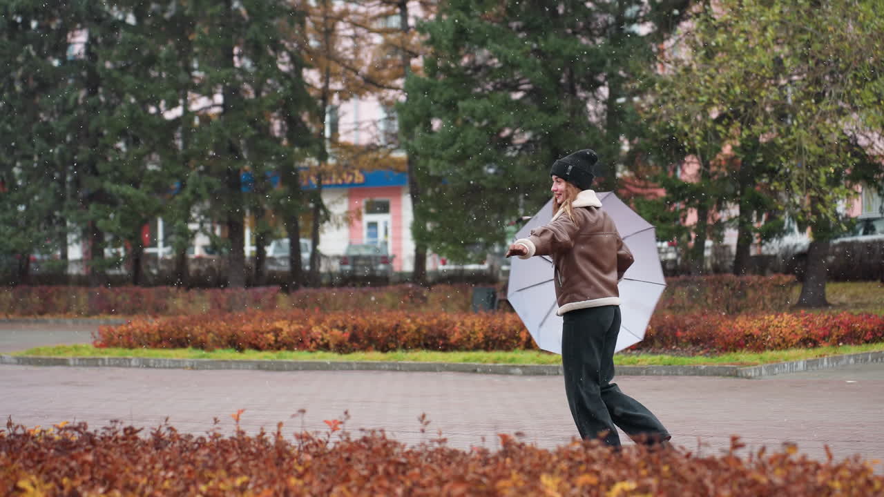 Female smiling, holding umbrella, wearing knit cap, brown shearling jacket, black trousers, happily turning around in light snowfall, enjoying winter day, spreading positive energy in cold weather