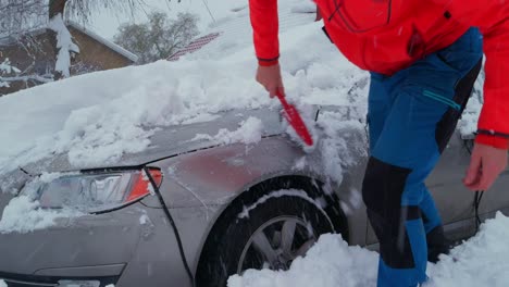 man uses snow brush to remove snow from car after blizzard