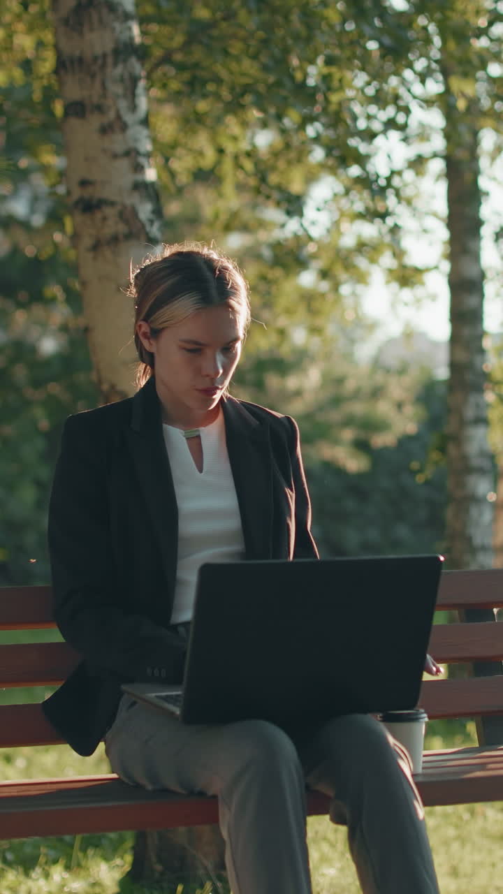 Designer in blazer concentrato su un progetto di design utilizzando un laptop, seduto in un parco all'aperto durante il giorno, con una tazza di caffè accanto, circondato da vegetazione e luce naturale