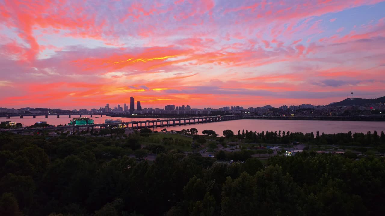 Aerial view of Seoul at sunset with vibrant purple and pink clouds, Banpo Bridge traffic over the Han River, Namsan Tower on the hill, and Hangang Park greenery in the foreground