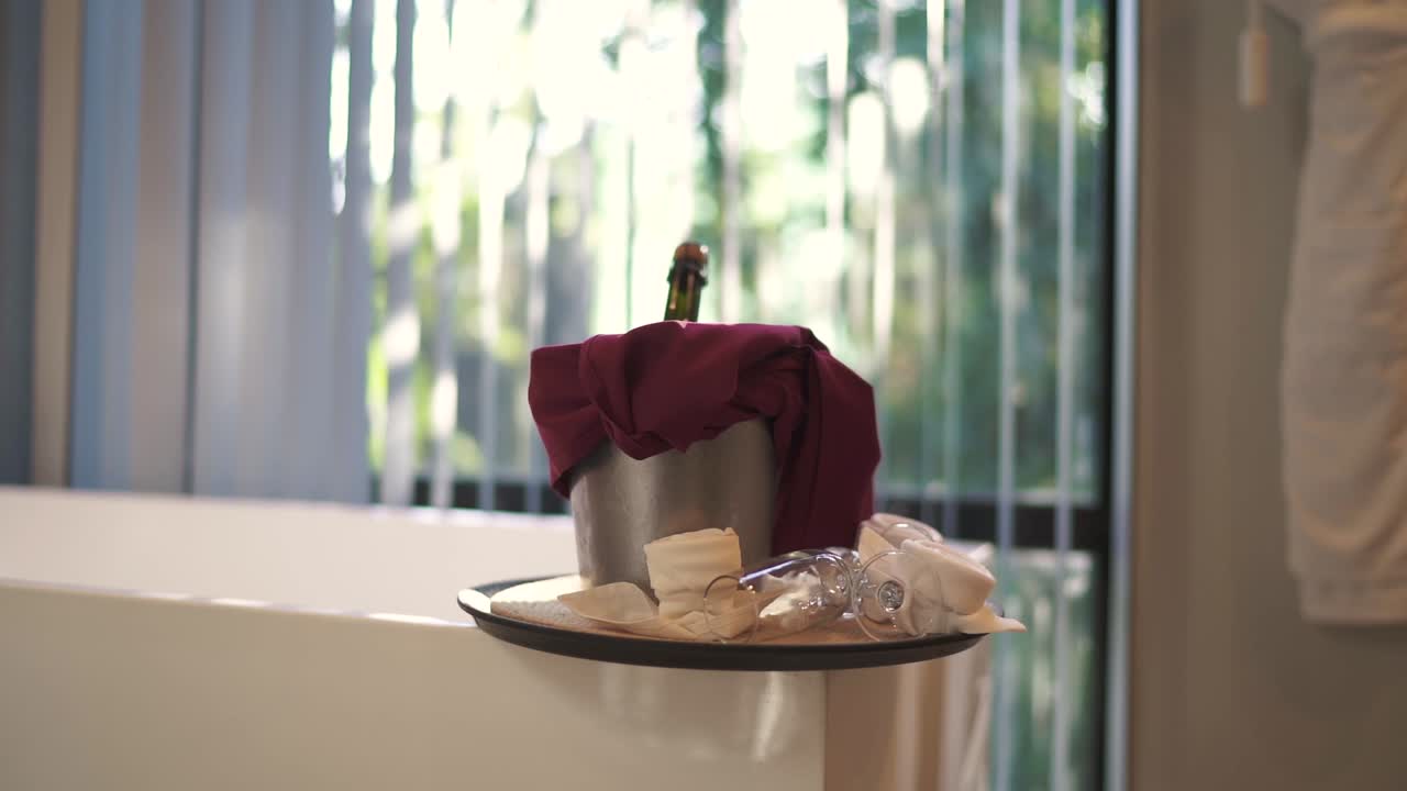 botella de champán enfriando en un cubo de hielo con dos vasos y pétalos en una bandeja cerca de una bañera en una habitación de hotel celebrando luna de miel o aniversario, concepto de escapada romántica, disparo en cámara lenta