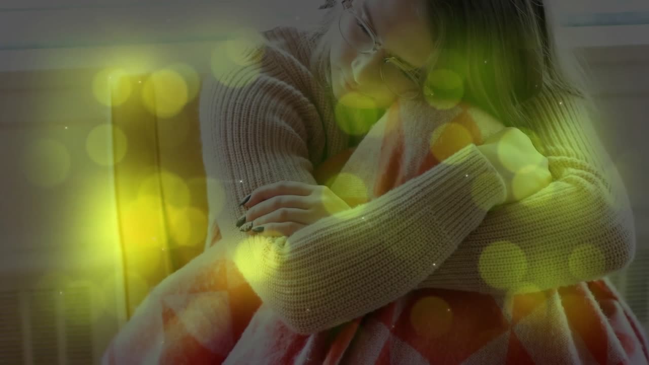 Donna accoccolata sul divano vicino alla finestra, che abbraccia una coperta, seguendo una sovrapposizione di bokeh giallo a sostegno del benessere.