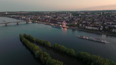 Mainzer-Sommernacht-Mit-Stadtlichtern-Und-Schiffen-Auf-Dem-Rhein-Vor-Orangefarbenem-Himmel-Durch-Eine-Filmische-Drohnenaufnahme