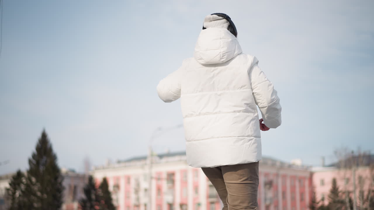 Round view student wearing white puffer coat, beanie and headphones dancing on snowy tiled square with blurred billboard and pastel buildings in urban winter plaza under clear blue sky