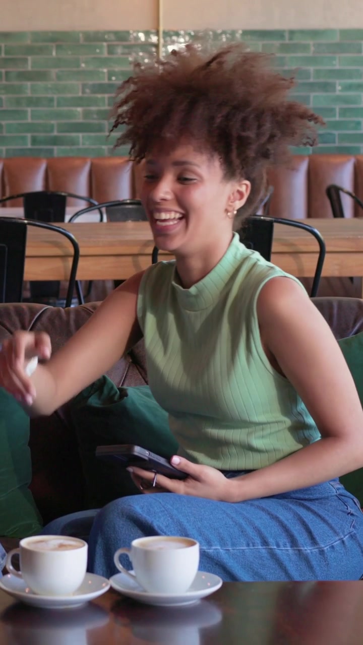 Video verticale: Donna con top color menta che legge e tocca il telefono, mostrandolo a un'amica al bar, ridendo.