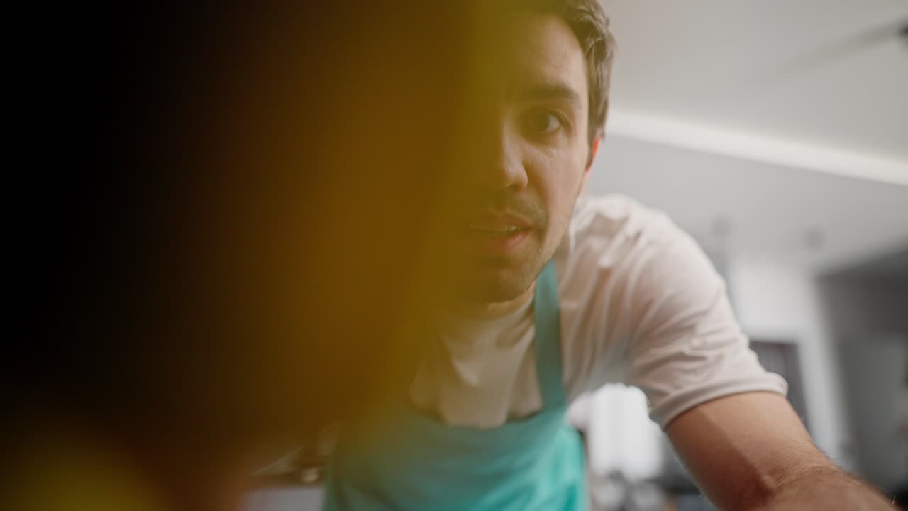 Confident brunette guy with stubble in a white T-shirt and blue apron wipes the camera with a yellow rag and poses with it while cleaning the kitchen in a modern apartment