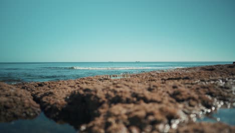 Costa-Oceánica-Plana,-Con-Barcos-De-Carga-Y-Olas-Que-Golpean-Las-Rocas-Suavemente-Bajo-La-Luz-Del-Sol-Gran-Angular-4k