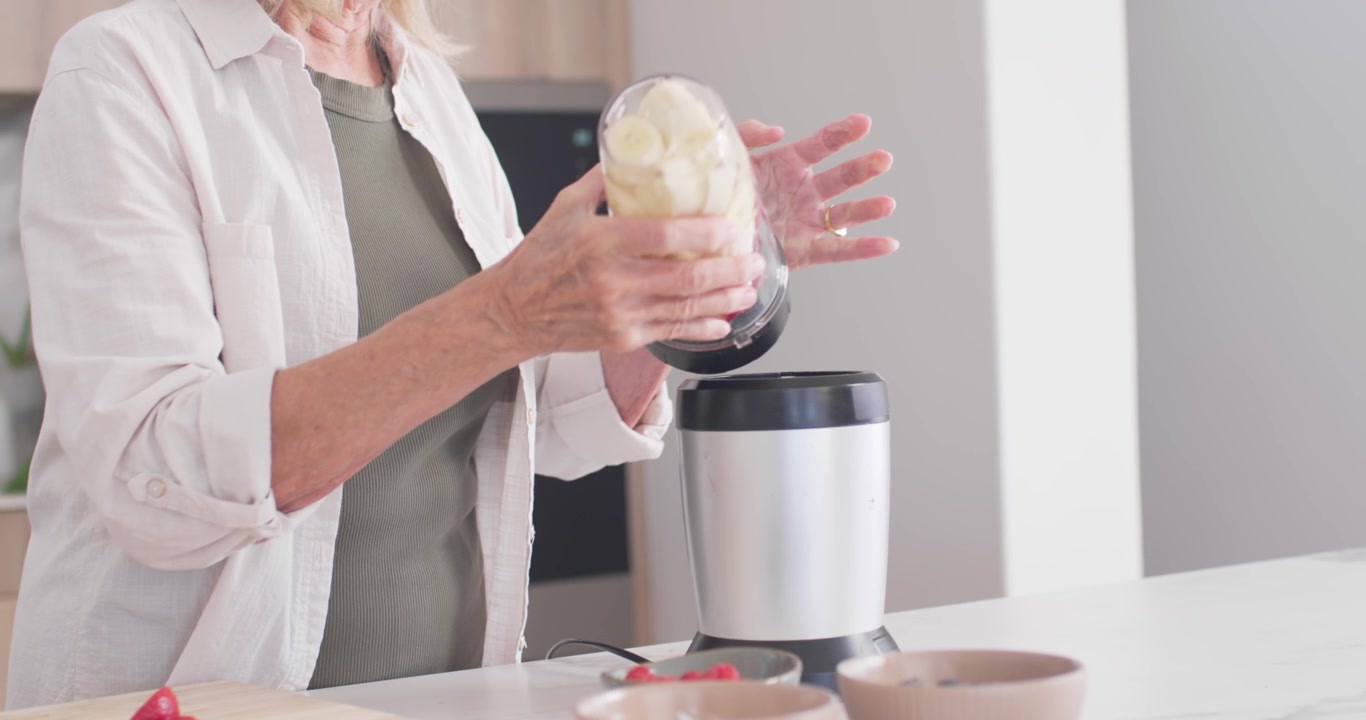 Donna anziana che tiene una tazza del frullatore con frutta, fissa il coperchio e capovolge la tazza sulla base del frullatore sul bancone.