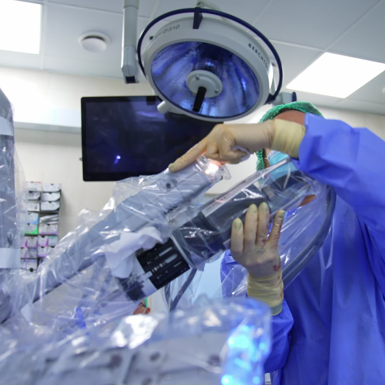 Medic rotates the robotic arm of Da Vinci system covered in plastic. Specialist looks at the screen in front of her in the modern surgery room