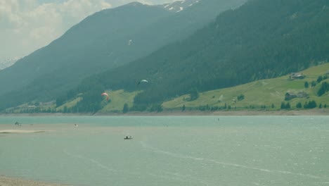 Toma-Completa,-Vista-Panorámica-De-La-Gente-Haciendo-Kitesurf-En-Medio-De-Reschensee-En-Italia,-Un-Día-Soleado-En-El-Fondo